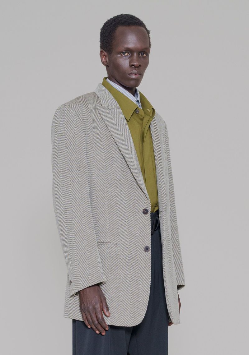 A dark-skinned male fashion model poses in a studio wearing a textured blazer, olive green shirt, and black slacks, against a grey backdrop.