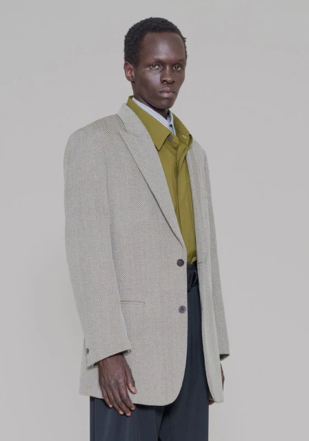 A dark-skinned male fashion model poses in a studio wearing a textured blazer, olive green shirt, and black slacks, against a grey backdrop.