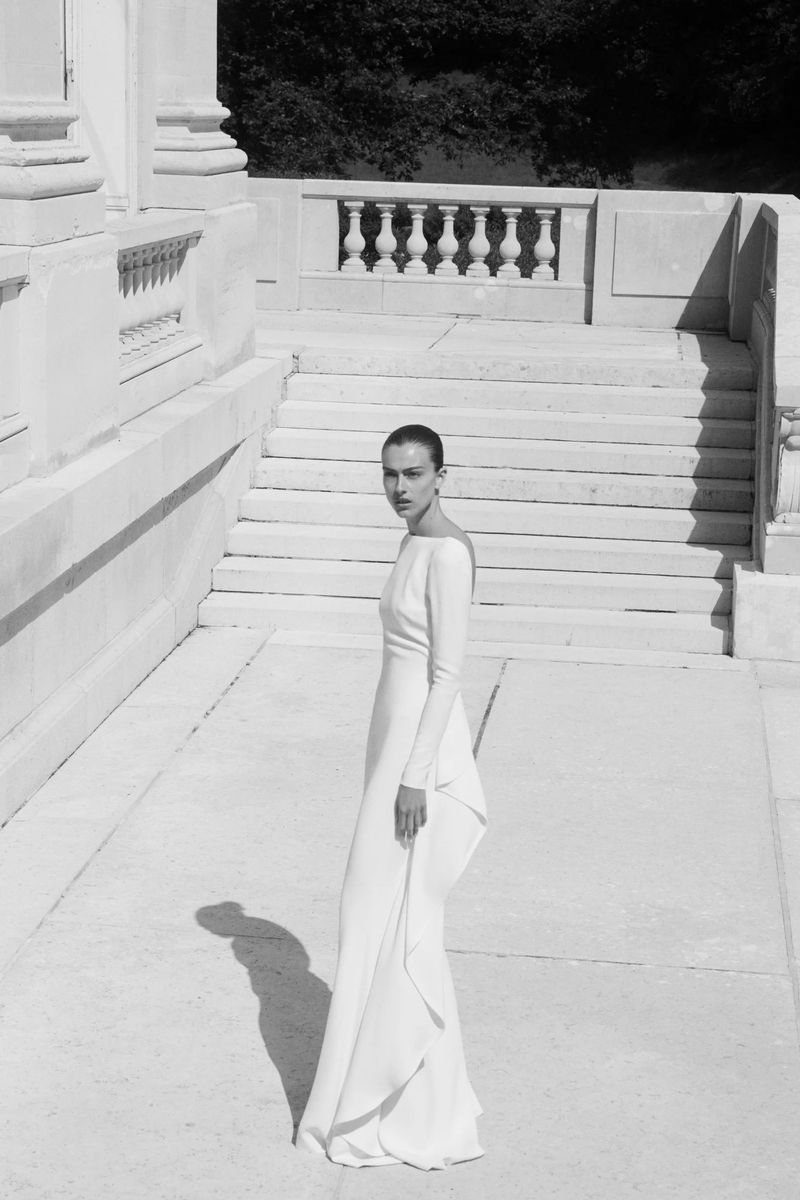 Woman in a long-sleeve white gown stands on stone steps near a building with pillars, gazing back over her shoulder.