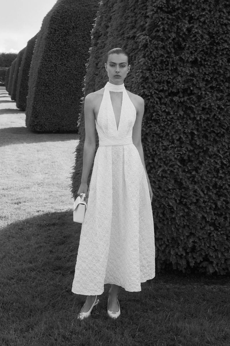 Elegant adult female model poses in a white halter dress with a white handbag, standing outdoors in a garden setting with trimmed hedges.