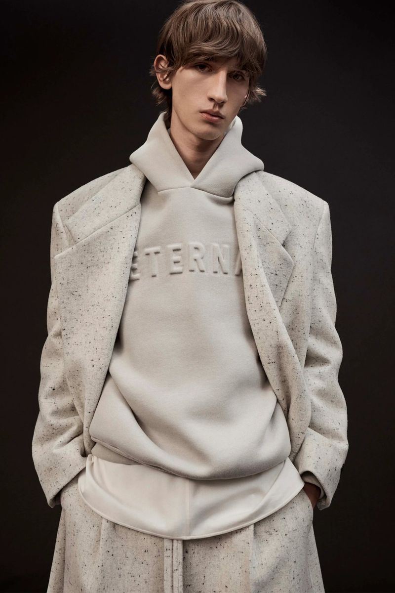 Fashion shot of male model in a speckled coat, hoodie and pants against a dark grey backdrop in a studio. The hoodie reads 'Eternal'.