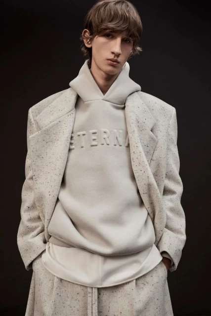 Fashion shot of male model in a speckled coat, hoodie and pants against a dark grey backdrop in a studio. The hoodie reads 'Eternal'.
