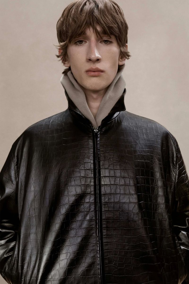 Portrait of a young adult man wearing a black crocodile-textured jacket with a gray hoodie, shot in a photo studio with soft lighting.
