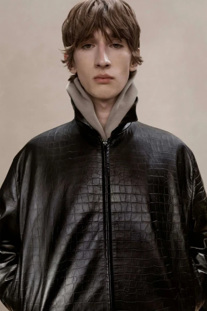 Portrait of a young adult man wearing a black crocodile-textured jacket with a gray hoodie, shot in a photo studio with soft lighting.
