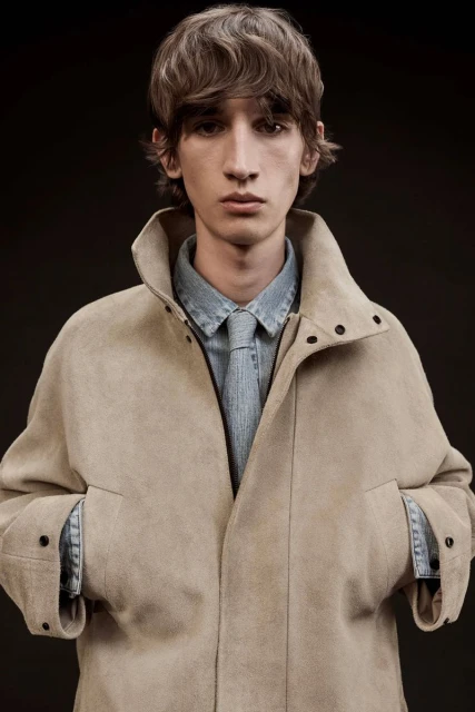 Portrait of a young adult man in a light brown coat and denim shirt with tie, medium shot, against a dark neutral background.