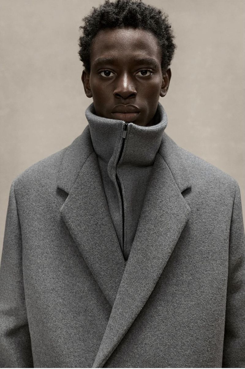 Portrait of a dark-skinned adult man wearing a grey coat and turtleneck indoors against a neutral backdrop in a medium shot.