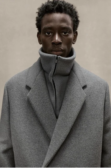 Portrait of a dark-skinned adult man wearing a grey coat and turtleneck indoors against a neutral backdrop in a medium shot.