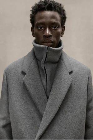 Portrait of a dark-skinned adult man wearing a grey coat and turtleneck indoors against a neutral backdrop in a medium shot.