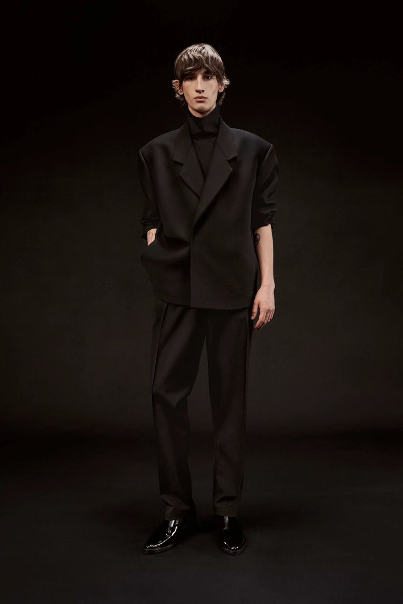 Model wearing an all-black oversized tailored suit and patent leather dress shoes standing against a black studio backdrop.