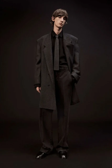 Adult male fashion model in a dark grey overcoat, grey suit, and black patent-leather shoes posing in a photo studio with a dark background.