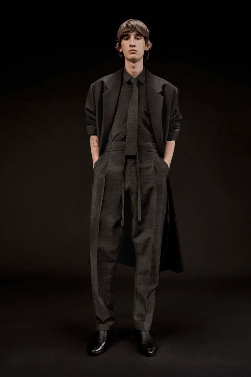 A slender man is posing in a dark grey suit and black overcoat in a studio setting. Full-length shot with neutral expression.