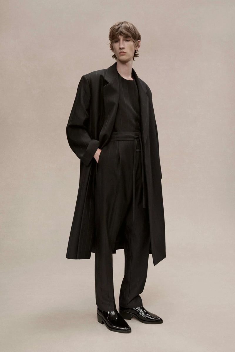 A fair-skinned man with dark hair poses in a black long-sleeved shirt, wide-leg pants, a long coat, and patent leather shoes in a studio.
