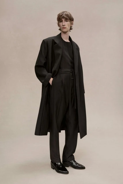 A fair-skinned man with dark hair poses in a black long-sleeved shirt, wide-leg pants, a long coat, and patent leather shoes in a studio.
