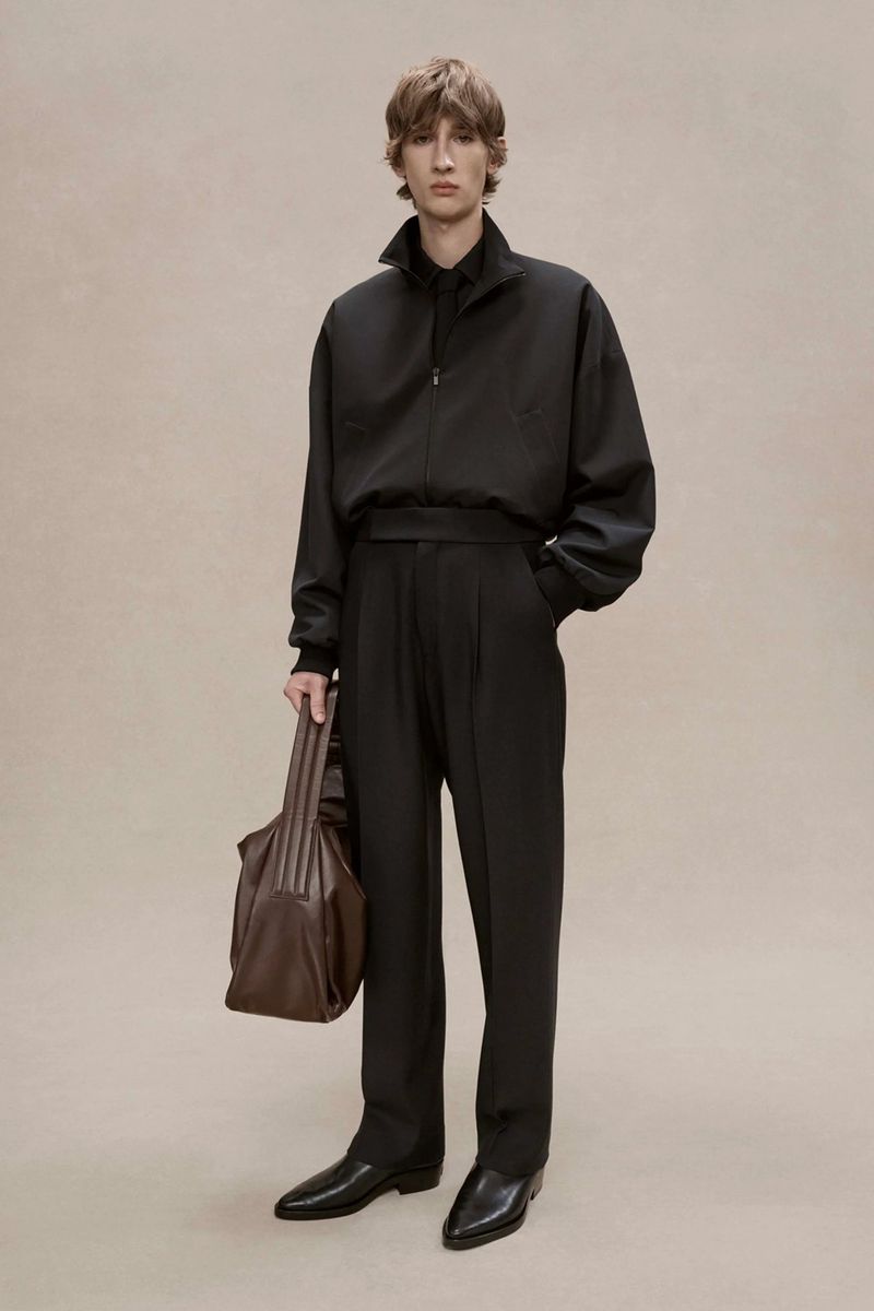 A stylish adult male model poses in a black jacket, black trousers, and black shoes while holding a brown leather bag. Studio setting.