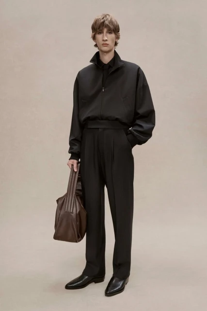 A stylish adult male model poses in a black jacket, black trousers, and black shoes while holding a brown leather bag. Studio setting.