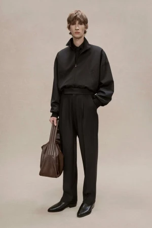 A stylish adult male model poses in a black jacket, black trousers, and black shoes while holding a brown leather bag. Studio setting.