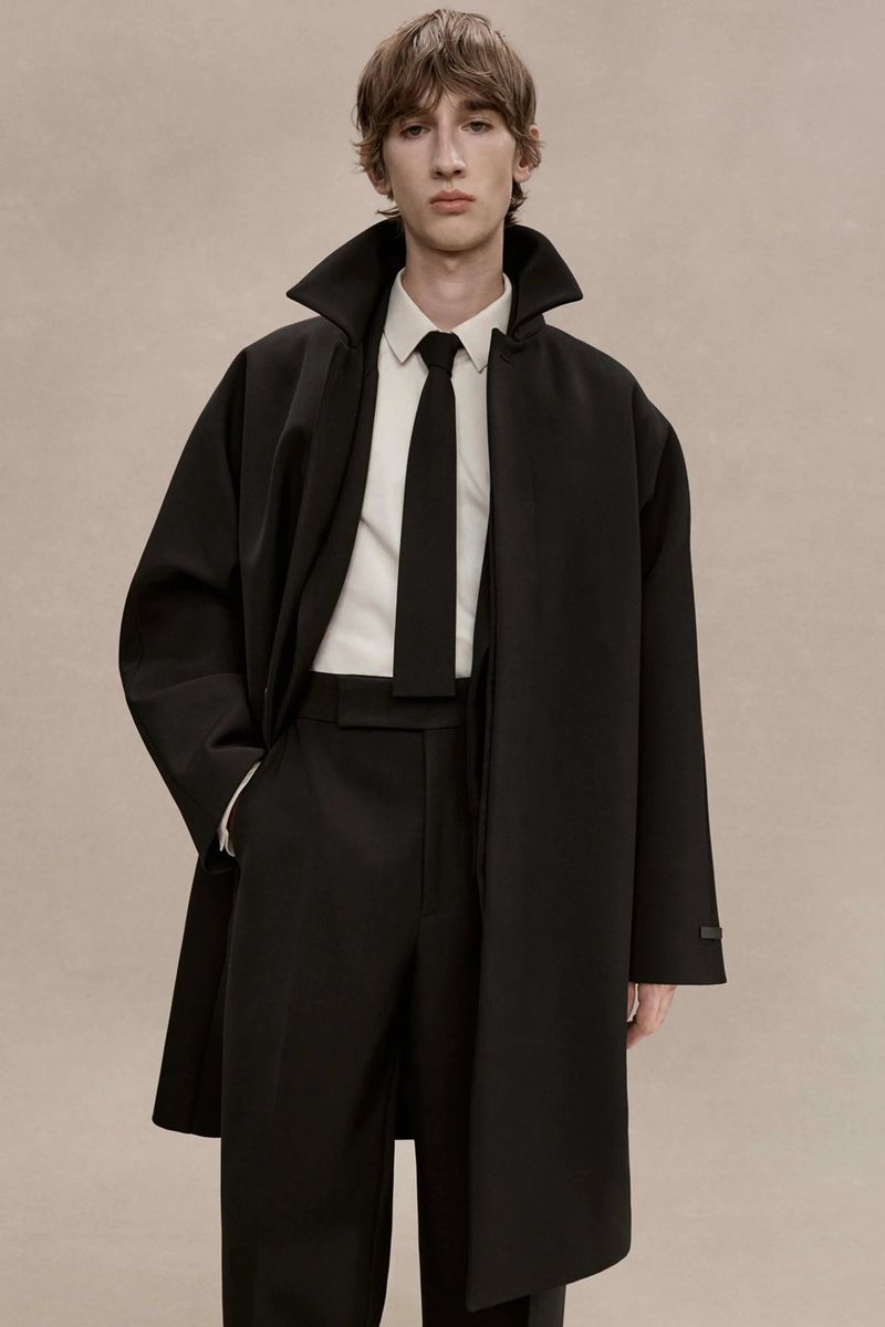 Model wearing an oversized black coat with a popped collar, white shirt, and black tie against a neutral studio backdrop.