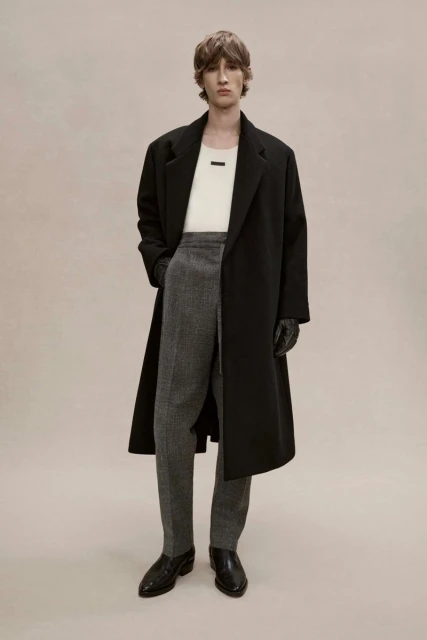 Male model in a long black coat, ivory top, and grey patterned trousers stands against a light background in a fashion editorial.