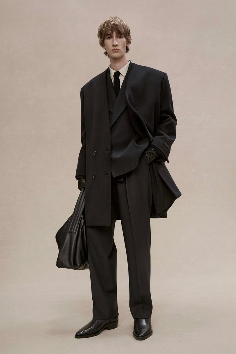 Male fashion model posing in an oversized black suit, black leather shoes, and carrying a matching tote bag against a neutral backdrop.