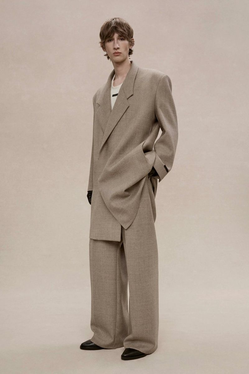 Male model wearing an oversized taupe wool suit and wide-leg trousers posing against a seamless warm neutral studio backdrop.