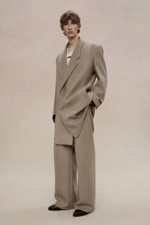 Male model wearing an oversized taupe wool suit and wide-leg trousers posing against a seamless warm neutral studio backdrop.