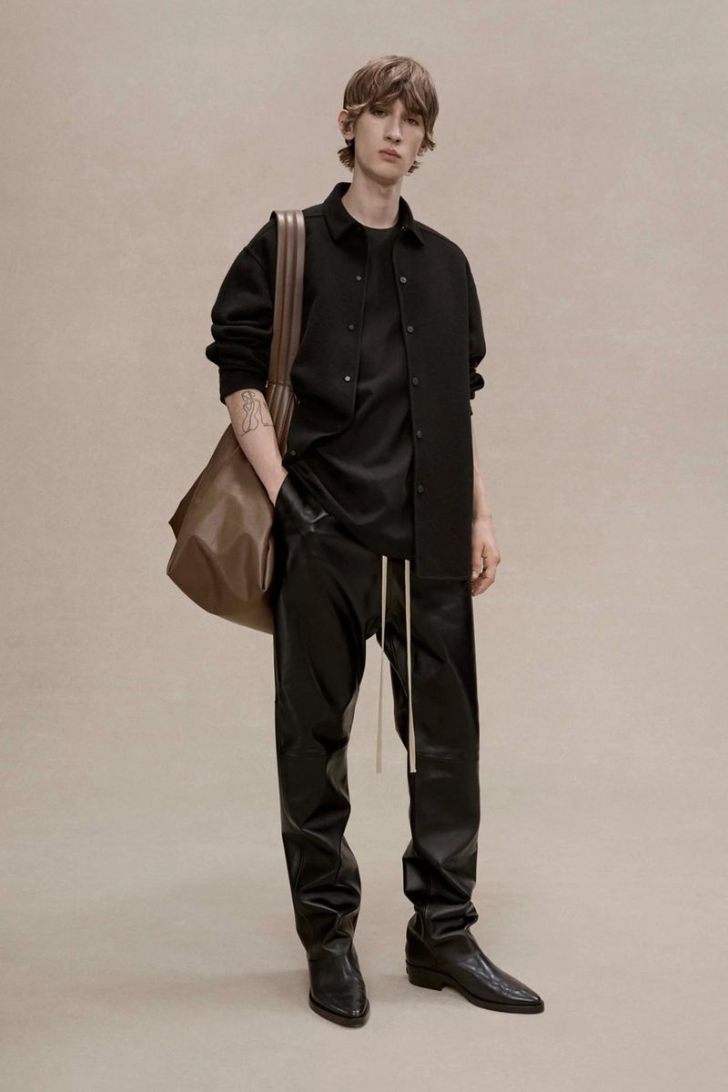 A slender adult male fashion model is posing indoors wearing a black top, jacket, leather trousers, boots, and holding a leather bag.