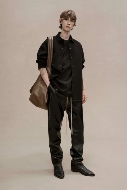 A slender adult male fashion model is posing indoors wearing a black top, jacket, leather trousers, boots, and holding a leather bag.