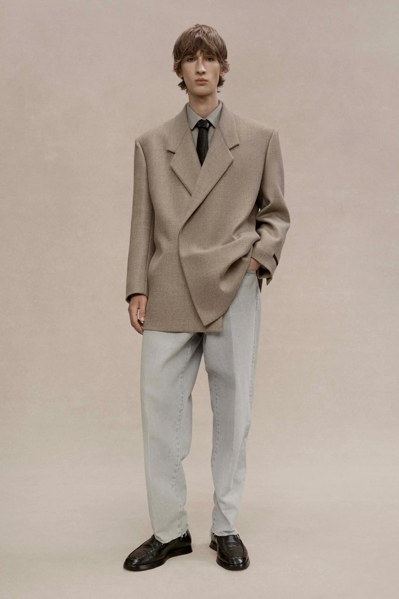 Male fashion model in a tan blazer, grey dress shirt, black tie, and light grey pants poses in a studio with neutral background.