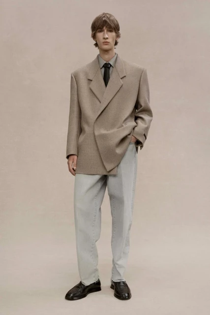 Male fashion model in a tan blazer, grey dress shirt, black tie, and light grey pants poses in a studio with neutral background.