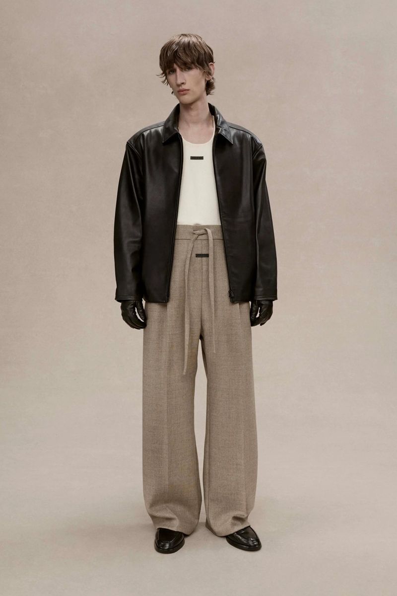 Full-length shot of a male fashion model wearing a black leather jacket, cream top, and pleated beige trousers, posing in a studio.