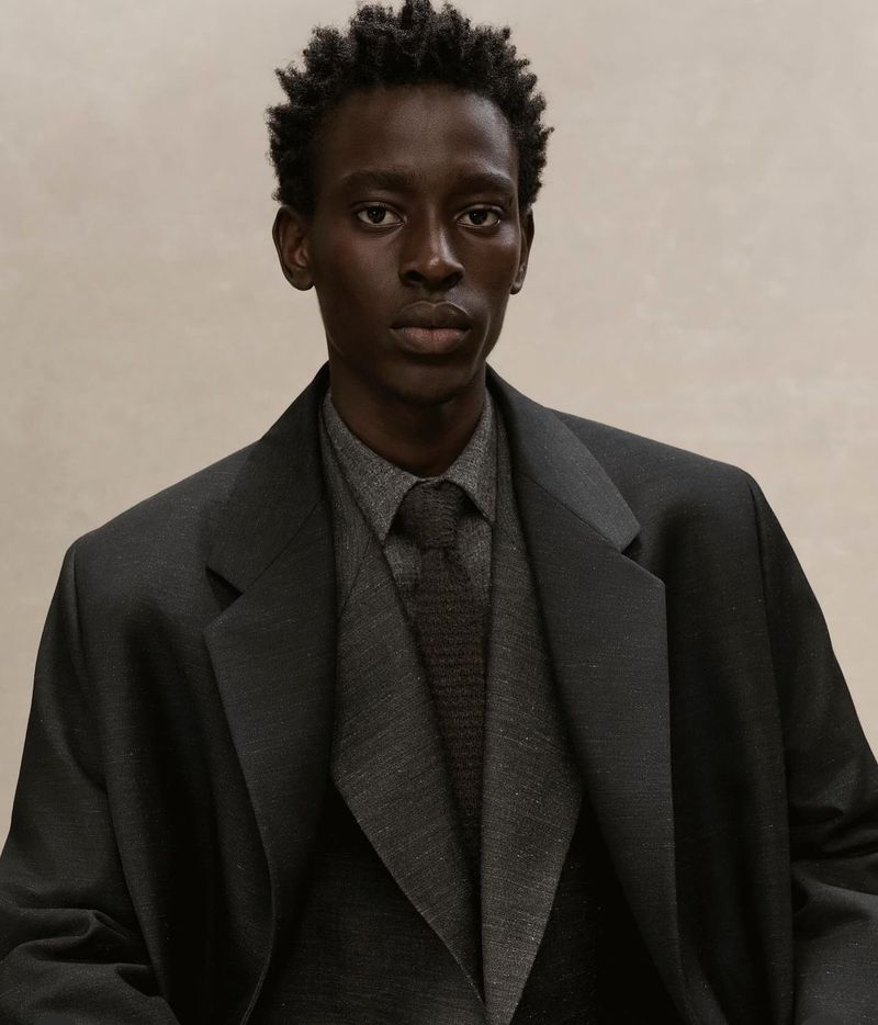 Dark-skinned adult man in a grey collared shirt, dark grey tie, and charcoal blazer against a neutral background in a studio portrait.