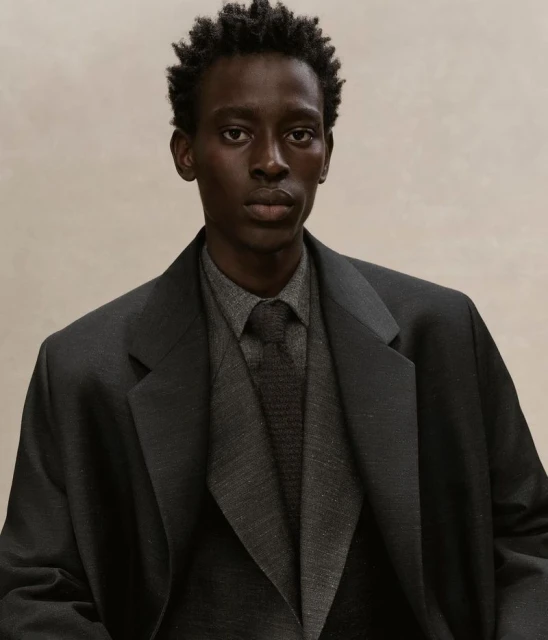 Dark-skinned adult man in a grey collared shirt, dark grey tie, and charcoal blazer against a neutral background in a studio portrait.