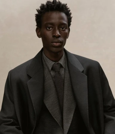 Dark-skinned adult man in a grey collared shirt, dark grey tie, and charcoal blazer against a neutral background in a studio portrait.