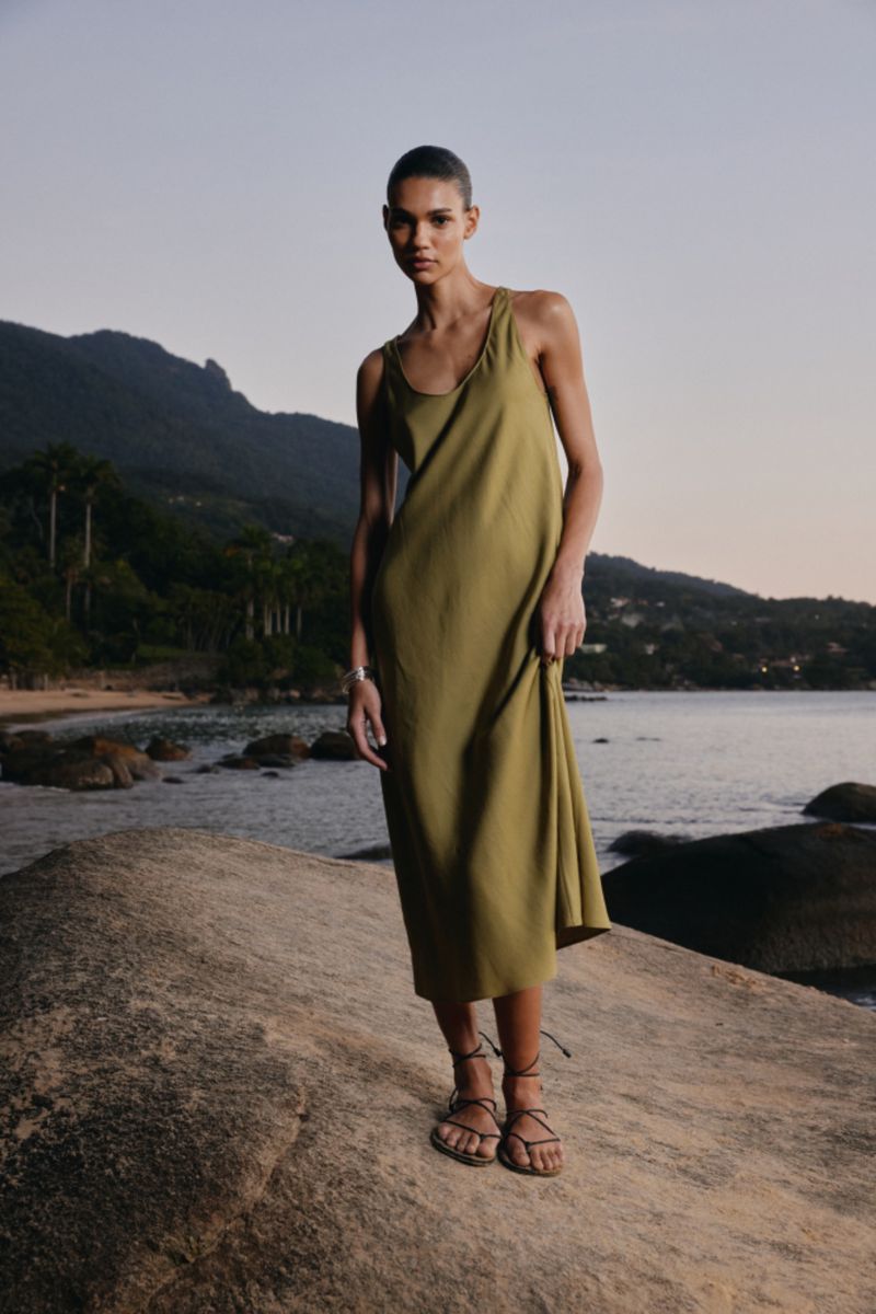 A woman in a tan dress stands on a rock formation by the sea. Mountain silhouettes in the background, creating a serene fashion shot.