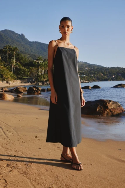 A fair-skinned woman in a black dress poses on a sandy beach with mountains and ocean in the background, enjoying the sunny weather.