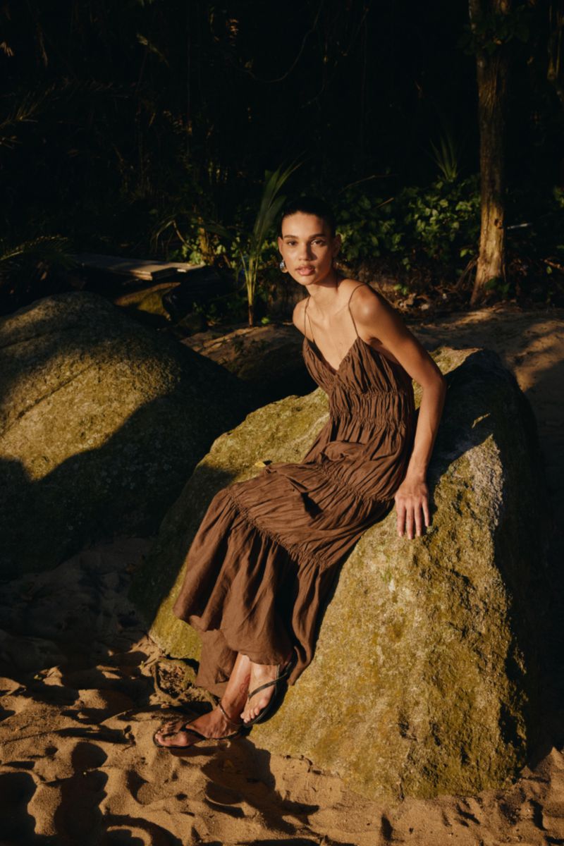 A woman in a brown dress sits on a large rock in an outdoor setting with sand, wearing flat sandals and gazing directly at the camera.