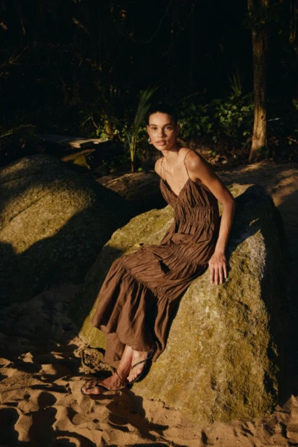 A woman in a brown dress sits on a large rock in an outdoor setting with sand, wearing flat sandals and gazing directly at the camera.