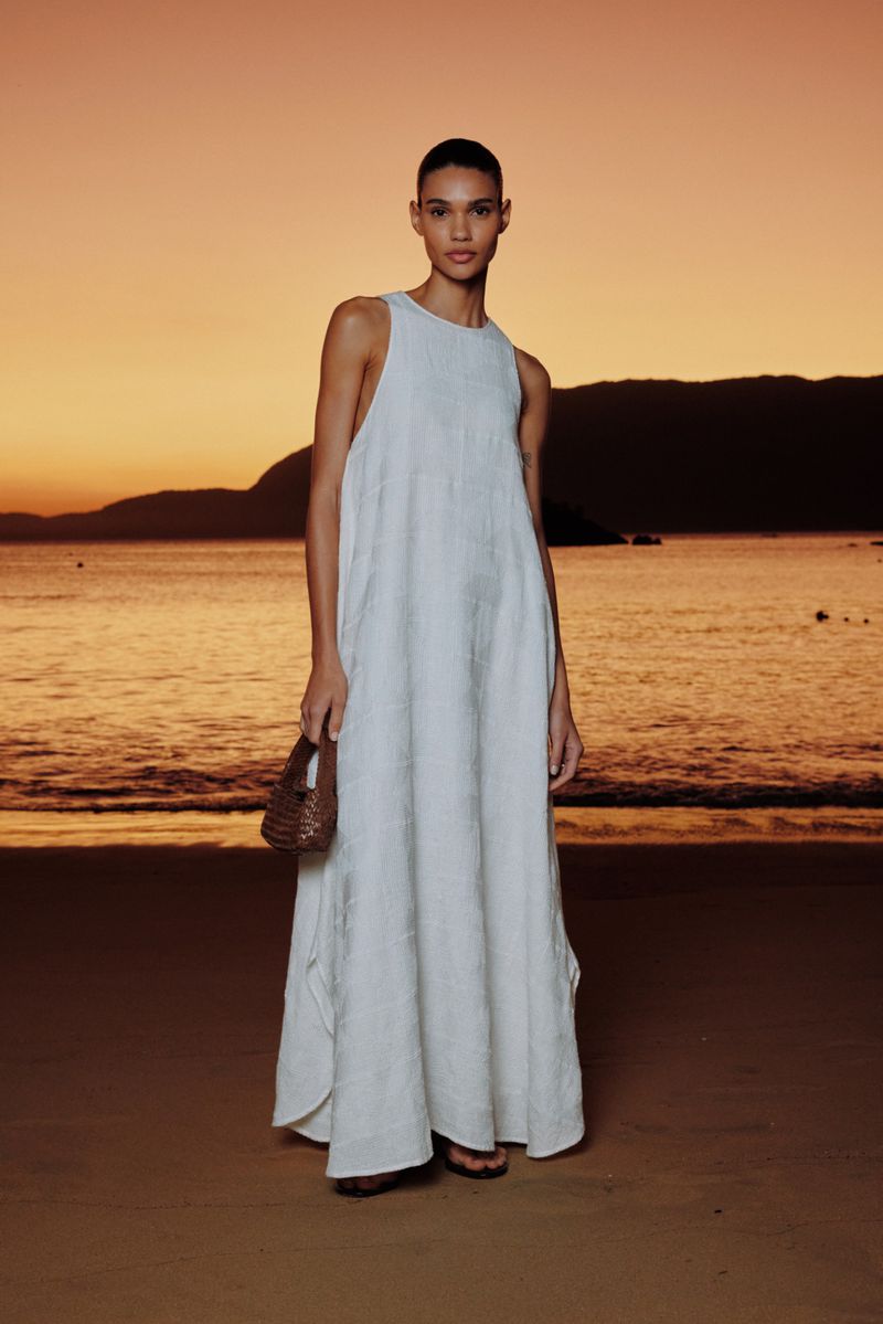 A woman in a loose white dress stands on a sandy beach at sunset, holding a woven bag, with ocean and orange sky in the background.