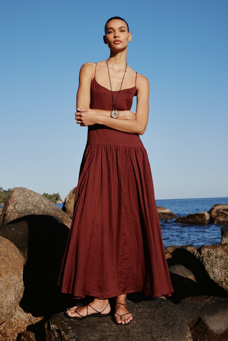 Woman stands on rocks near the sea wearing a maroon dress and pendant necklace, posing with arms crossed on a sunny day.