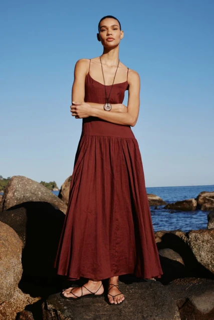 Woman stands on rocks near the sea wearing a maroon dress and pendant necklace, posing with arms crossed on a sunny day.