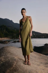 Model Barbara Valente posing on a coastal rock wearing an olive green sleeveless midi dress and strappy flat sandals.