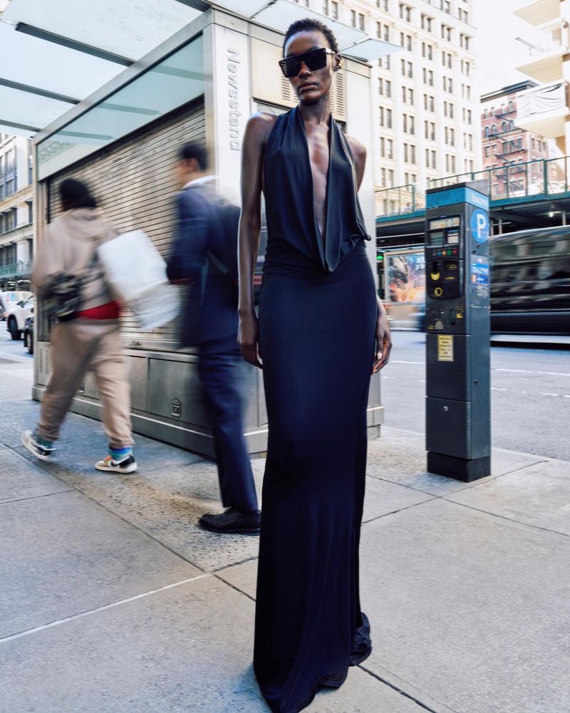 Woman in elegant black halter dress stands on city sidewalk, pedestrians and buildings in background, sunglasses, fashion editorial.