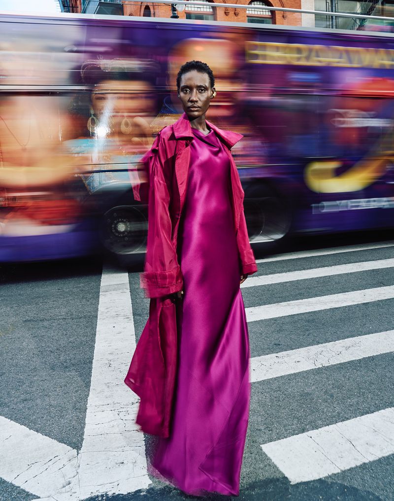 Adult female poses in a magenta dress and a red coat on a crosswalk with a blurred bus passing behind her in an urban setting.