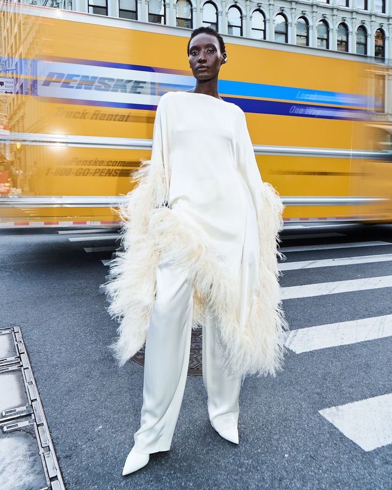 A fashion shot features a single, elegant Black woman standing on a city street wearing a flowing white dress with feathered sleeves.