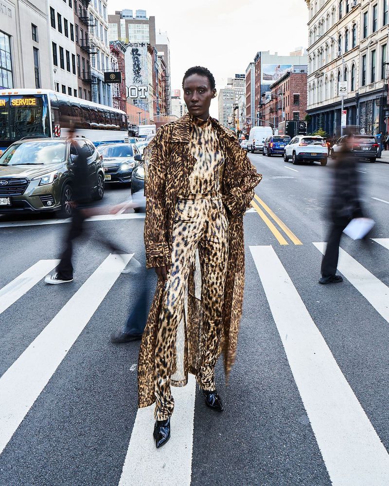 A woman in a leopard-print outfit stands on a crosswalk in a city street with blurred motion from other pedestrians and traffic around her.