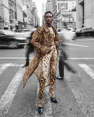 An African American woman in a leopard print coat and jumpsuit poses on a crosswalk in a city street, with cars and people blurred in motion.