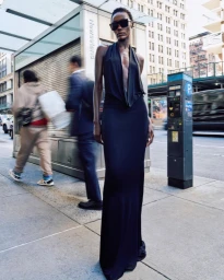 Model in a black halter gown and sunglasses standing on a city sidewalk with blurred pedestrians