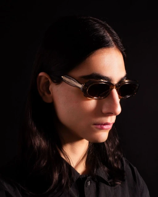 Close-up studio shot of an adult with dark hair, wearing brown-framed sunglasses and a black shirt against a solid black background.