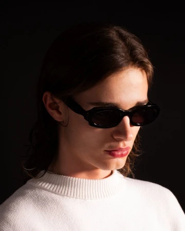 Model wearing chunky black sunglasses and a white crewneck knit sweater against a dark black background.
