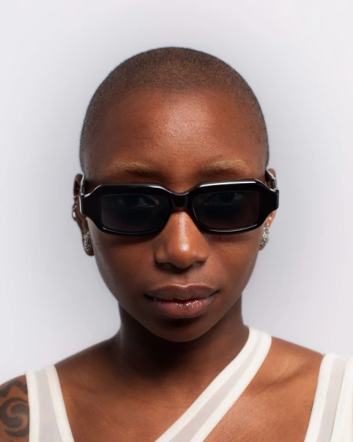 Headshot of a dark-skinned, bald adult female with black sunglasses and earring against a solid white studio backdrop.